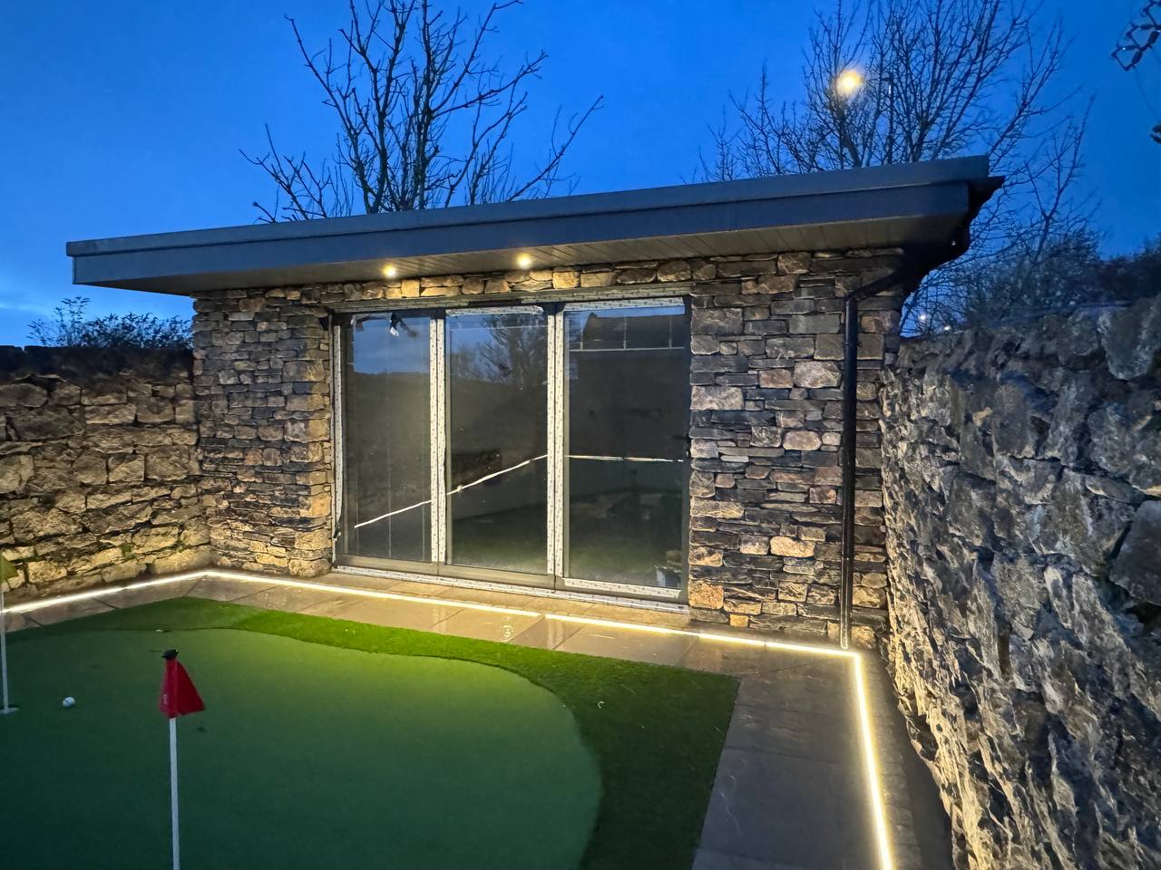 High-end garden room with natural stone walling, glass frontage, lighting and putting green by Sam Shaw Kilburn Stonework Ltd